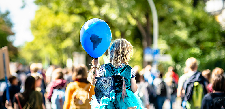 Eine protestierende Menschenmenge im Hintergrund, die unscharf dargestellt wird. Im Vordergrund ist ein kleines Mädchen fokussiert, das einen blauen Luftballon trägt, auf dem die Erde abgebildet wird.